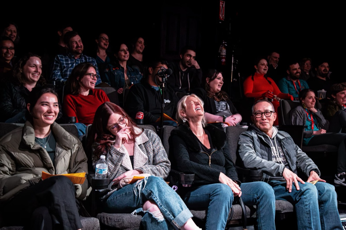 A packed theatre audience mid-laugh, rows of people watching the stage.