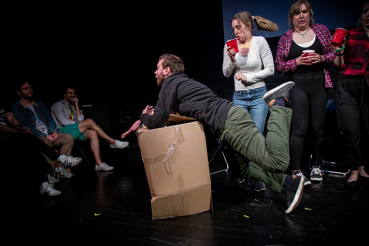 A chaotic party scene — one performer sprawled over a cardboard box while four others look on.