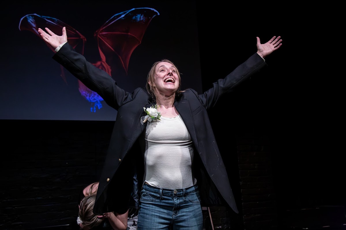 A performer on a dark stage with both arms thrown wide, mid-sketch.