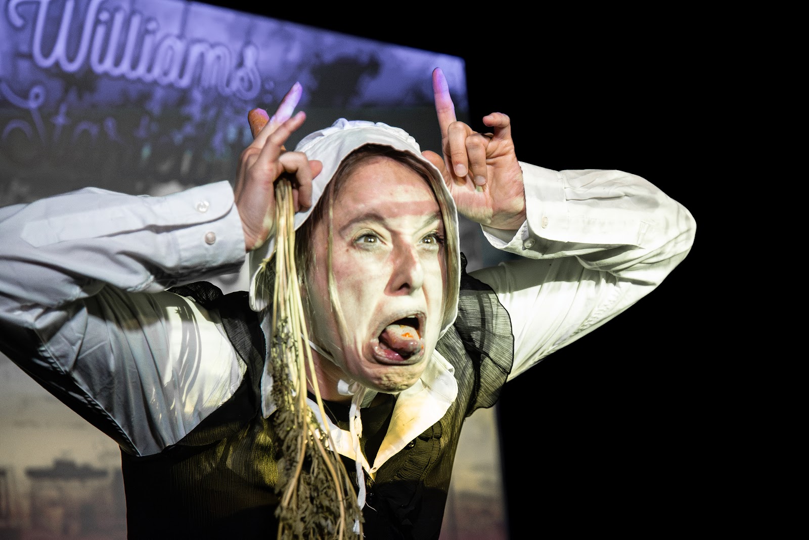 A performer mid-scream on a dark stage, hands raised beside her head.